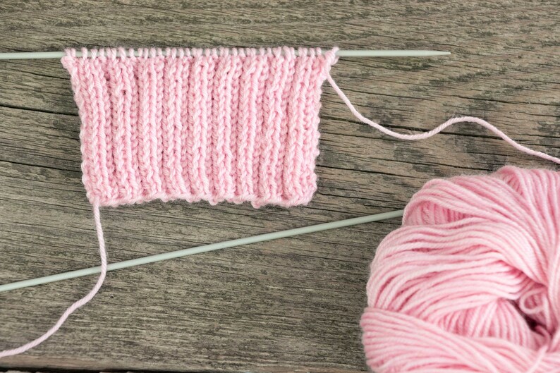 Puede incluir: Un ovillo de hilo rosa con una muestra de tejido de punto rosa en agujas de tejer. La muestra est&aacute; sobre una superficie de madera.