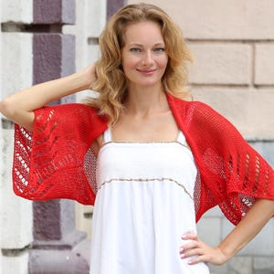 K&ouml;nnte beinhalten: Ein leuchtend roter Strick-Bolero mit einem durchbrochenen Spitzenmuster an den &Auml;rmeln und einem glatt gestrickten R&uuml;cken. Das Kleidungsst&uuml;ck hat eine leichte Textur und enth&auml;lt den Text HITS KNITS, der dezent in den linken &Auml;rmel eingearbeitet ist.