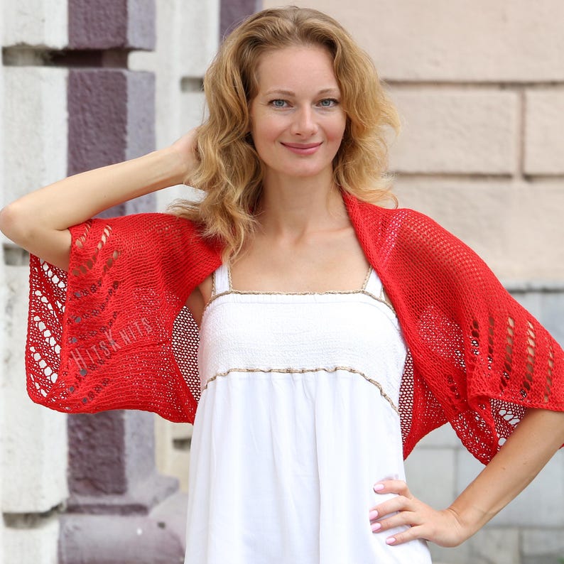 K&ouml;nnte beinhalten: Ein leuchtend roter Strick-Bolero mit einem durchbrochenen Spitzenmuster an den &Auml;rmeln und einem glatt gestrickten R&uuml;cken. Das Kleidungsst&uuml;ck hat eine leichte Textur und enth&auml;lt den Text HITS KNITS, der dezent in den linken &Auml;rmel eingearbeitet ist.