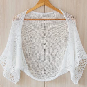 May include: A white, open-front knitted shawl with short sleeves, displayed on a wooden hanger. The shawl has a delicate, open weave design, with a slightly scalloped edge on the sleeves. The garment is made of a soft, lightweight material.