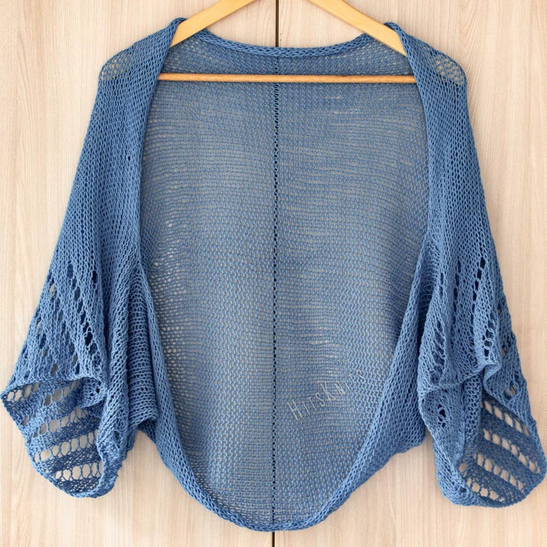 K&ouml;nnte beinhalten: Ein blauer Bolero mit einer offenen Strickkonstruktion und einer zarten, luftigen Textur. Das Kleidungsst&uuml;ck weist eine mittlere vertikale Naht und komplizierte, weiter auseinanderliegende Strickmuster an den &Auml;rmeln auf. Der Text HitsKnits ist sichtbar.