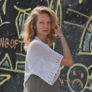May include: A white, open-weave knit shawl with decorative edging. The shawl is worn over a neutral-colored top. The background features graffiti art with yellow and black colors. The text "Hits Knits" is visible.