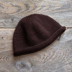 May include: A brown knitted beanie hat with a folded brim. The hat is lying on a wooden surface.