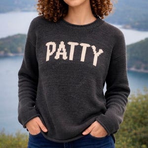 May include: Dark grey knitted sweater with the name "PATTY" in large, white block letters across the chest. The sweater has a crew neck and long sleeves. The background shows a blurred view of a body of water and green hills.