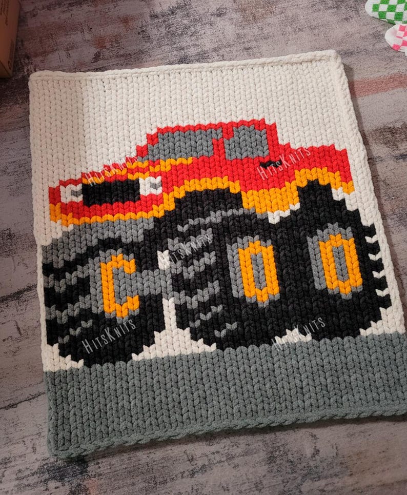 May include: A handmade, square, crocheted blanket featuring a pixelated monster truck design. The truck is red, orange, yellow, gray, and black, with the word "COO" on the tire. The blanket has a white top, gray bottom, and the text "HitsKnits".