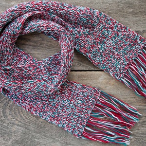 May include: A red, white, and green knitted scarf with fringe on both ends. The scarf is folded in half and laid on a wooden surface.