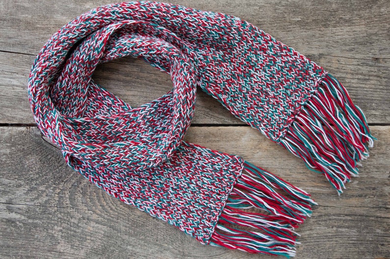 May include: A red, white, and green knitted scarf with fringe on both ends. The scarf is folded in half and laid on a wooden surface.