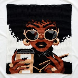 May include: Crocheted square depicting a woman with sunglasses, holding a coffee cup and a cigarette. The design uses black, brown, tan, white, and gold. The text "Hits Knits" is visible. The piece has a modern, artistic style.