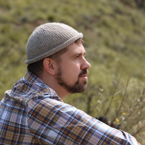 May include: A man wearing a gray knitted beanie and a plaid shirt is looking off to the side. He is standing in a field of green grass.