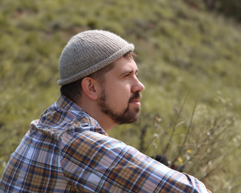 May include: A man wearing a gray knitted beanie and a plaid shirt is looking off to the side. He is standing in a field of green grass.