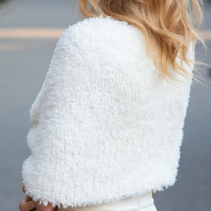 May include: A white, cropped shrug featuring a textured, shaggy faux fur finish. The garment has short sleeves and a rounded neckline, providing a soft, fuzzy appearance that covers the shoulders and upper back area with a dense, uniform pile.