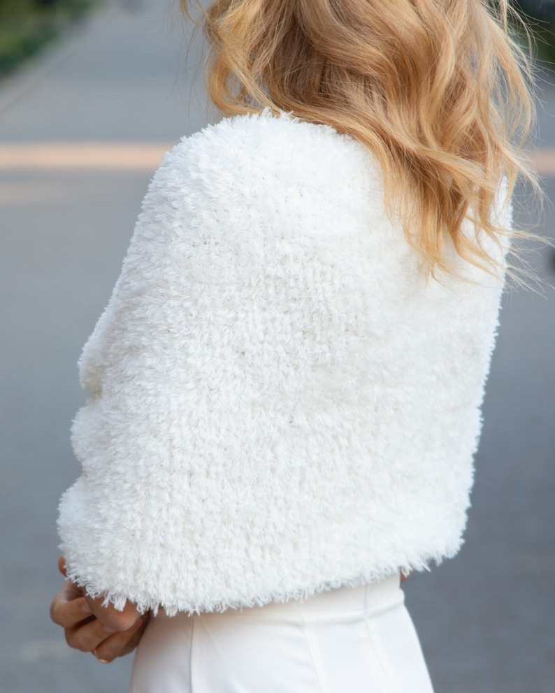 May include: A white, cropped shrug featuring a textured, shaggy faux fur finish. The garment has short sleeves and a rounded neckline, providing a soft, fuzzy appearance that covers the shoulders and upper back area with a dense, uniform pile.