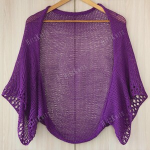 May include: A vibrant purple knitted shawl with a delicate openwork design. The shawl is draped over a wooden hanger, showcasing its flowing shape and intricate details. The edges feature a decorative pattern, adding a touch of elegance. The text "HitsKnits" is visible on the shawl.