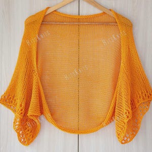 May include: A mustard yellow knitted shawl with a delicate open weave. The shawl is draped over a wooden hanger, showcasing its flowing design. The edges feature a decorative, openwork pattern. The text "HitsKnits" is visible on the shawl.