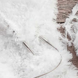 May include: A pair of silver circular knitting needles with a flexible cord, currently holding a project made of white fuzzy, textured yarn. A ball of the same white, soft, shaggy yarn is positioned to the side of the knitting needles.