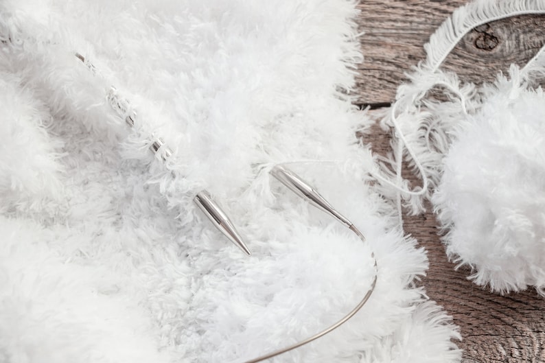 May include: A pair of silver circular knitting needles with a flexible cord, currently holding a project made of white fuzzy, textured yarn. A ball of the same white, soft, shaggy yarn is positioned to the side of the knitting needles.
