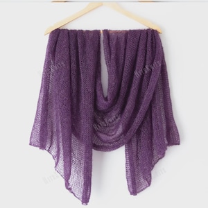 May include: A sheer, purple knitted shawl is displayed on a wooden hanger. The shawl is draped to show its texture and flow. The color is a deep, rich shade of purple, and the knit pattern is open and airy.