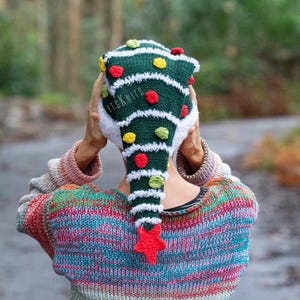 May include: A dark green knitted hat shaped like a Christmas tree with white fuzzy trim, decorated with small red, yellow, and light green pom-poms, and topped with a red star at the point. The text ITSKNITS is visible on the green fabric.