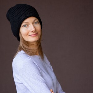 May include: A woman with long brown hair is wearing a black knit beanie and a light blue long-sleeved shirt. She is looking at the camera and smiling.