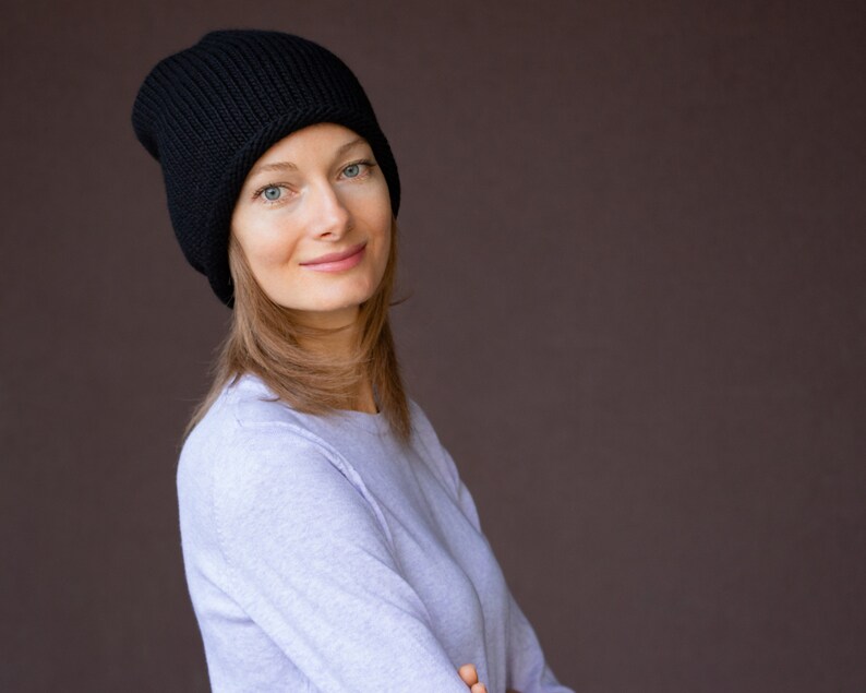 May include: A woman with long brown hair is wearing a black knit beanie and a light blue long-sleeved shirt. She is looking at the camera and smiling.
