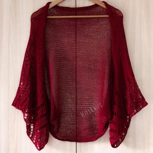 K&ouml;nnte beinhalten: Ein dunkelroter, vorne offener Bolero mit einer feinen, luftigen Strickstruktur am Hauptteil und einem komplexeren, durchbrochenen Spitzenmuster an den weiten &Auml;rmeln. Das Kleidungsst&uuml;ck enth&auml;lt den Schriftzug HitsKnits in passendem Rot.
