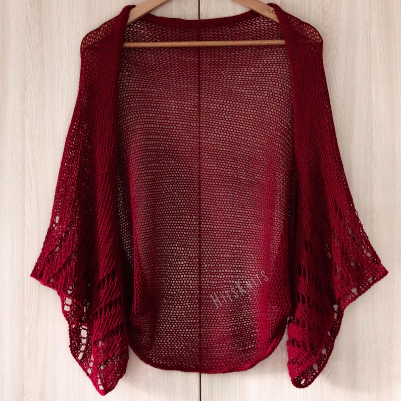 K&ouml;nnte beinhalten: Ein dunkelroter, vorne offener Bolero mit einer feinen, luftigen Strickstruktur am Hauptteil und einem komplexeren, durchbrochenen Spitzenmuster an den weiten &Auml;rmeln. Das Kleidungsst&uuml;ck enth&auml;lt den Schriftzug HitsKnits in passendem Rot.