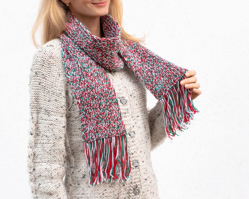 May include: A white, red, and green knitted scarf with fringe. The scarf is worn around the neck of a person wearing a white sweater.