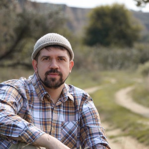 May include: A man wearing a beige knit beanie and a plaid shirt sits in a field with a dirt path behind him.