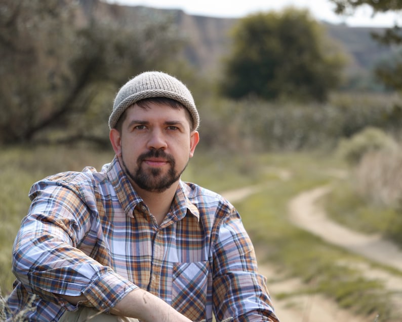 May include: A man wearing a beige knit beanie and a plaid shirt sits in a field with a dirt path behind him.