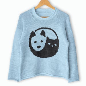 Può includere: Un maglione in maglia a maniche lunghe color azzurro con una grafica circolare sul davanti. Il design raffigura un cane bianco e un gatto nero disposti in uno stile yin-yang, con il cane a sinistra e il gatto a destra, entrambi rivolti in avanti.