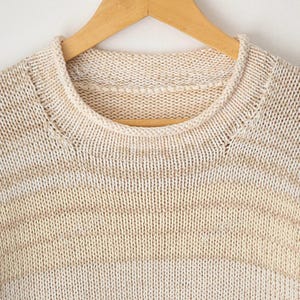May include: A crew neck sweater featuring a horizontal striped pattern in alternating shades of cream and light beige. The garment is crafted with a visible knit texture and is displayed on a wooden hanger, showcasing the round neckline.