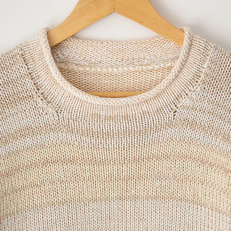 May include: A crew neck sweater featuring a horizontal striped pattern in alternating shades of cream and light beige. The garment is crafted with a visible knit texture and is displayed on a wooden hanger, showcasing the round neckline.