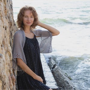 Peut inclure: Un châle tricoté gris clair avec des manches évasées. Le châle est porté sur une robe bleu foncé avec un petit motif blanc. La femme se tient près d'un mur de pierre et de la mer.