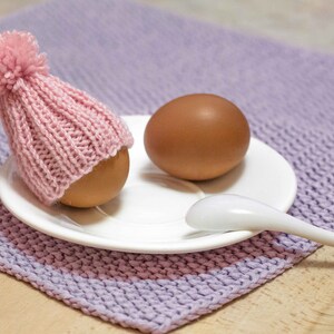 Puede incluir: Un huevo marr&oacute;n con un gorro de punto rosa con un pomp&oacute;n blanco est&aacute; sentado en un platillo blanco con una cuchara blanca a su lado. El platillo est&aacute; sobre una superficie de punto morado.
