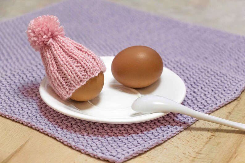 Puede incluir: Un huevo marr&oacute;n con un gorro de punto rosa con un pomp&oacute;n blanco est&aacute; sentado en un platillo blanco con una cuchara blanca a su lado. El platillo est&aacute; sobre una superficie de punto morado.