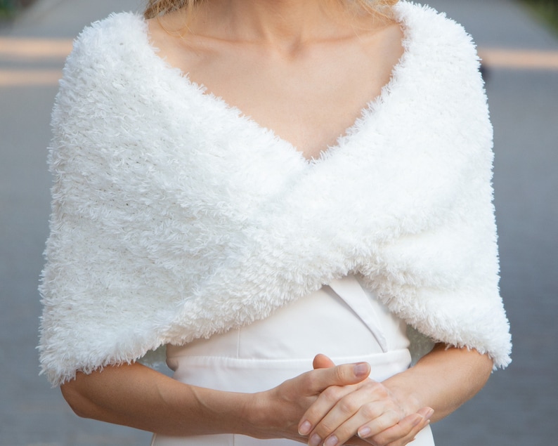 May include: A white faux fur shrug with a textured, shaggy appearance. The garment features a V-neckline and wraps across the chest, providing coverage over the shoulders and upper arms. The material has a soft, fuzzy pile throughout the piece.