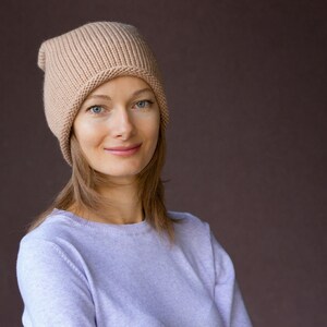 May include: A woman with long brown hair is wearing a light brown knitted beanie hat and a light purple sweater. She is looking at the camera and smiling.