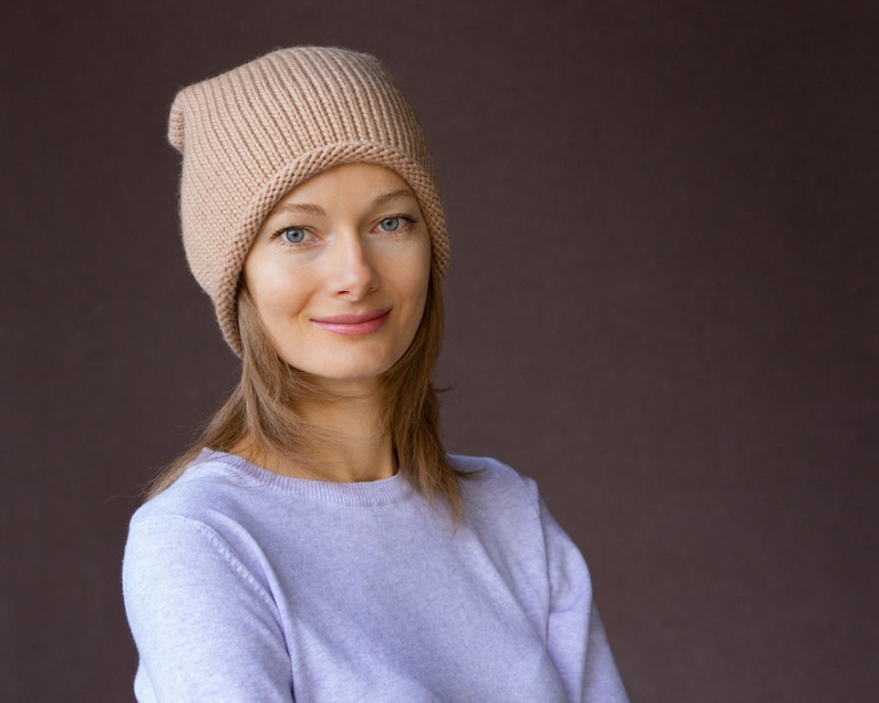 May include: A woman with long brown hair is wearing a light brown knitted beanie hat and a light purple sweater. She is looking at the camera and smiling.