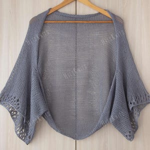 May include: A light gray knitted shawl with a delicate open weave design. The shawl is draped over a wooden hanger, showcasing its triangular shape and openwork edges. The garment is a soft, muted gray color.