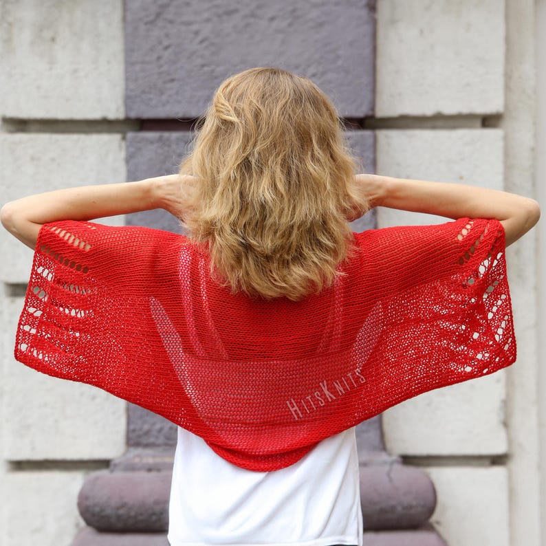 K&ouml;nnte beinhalten: Ein leuchtend roter Strickponcho mit einem durchbrochenen Spitzenmuster an den Seiten und einem glatt gestrickten Mittelteil. Das Kleidungsst&uuml;ck zeigt den Text HitsKnits in heller Schrift auf der unteren rechten Seite, getragen &uuml;ber einem wei&szlig;en Shirt.