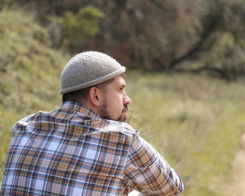 May include: A man wearing a gray knitted beanie and a plaid shirt is looking off to the side. He is standing in a grassy field.