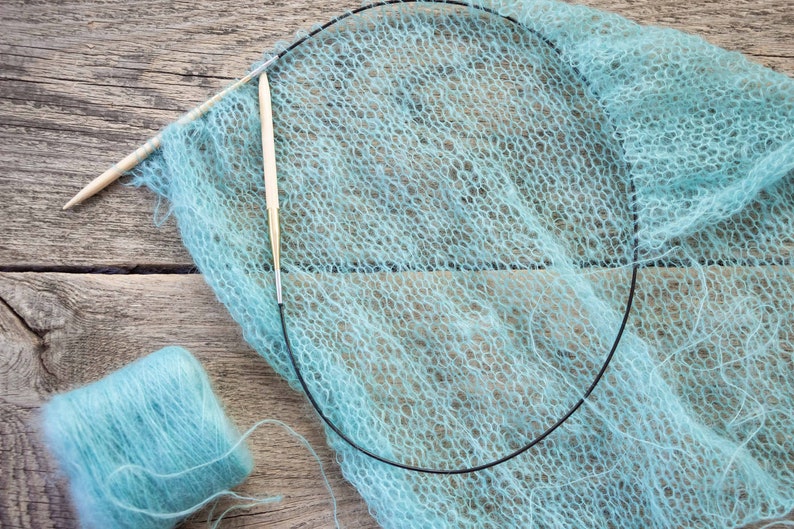 May include: A light blue yarn skein and a partially knitted scarf with two knitting needles. The scarf is made of a delicate, openwork stitch.