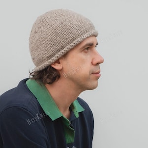 May include: A light beige, hand-knitted beanie with a rolled brim edge. The hat features a textured, chunky knit pattern in a neutral, speckled yarn. The word HitsKnits is visible as a watermark across the image, overlaying the hat and shirt.