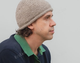 May include: A light beige, hand-knitted beanie with a rolled brim edge. The hat features a textured, chunky knit pattern in a neutral, speckled yarn. The word HitsKnits is visible as a watermark across the image, overlaying the hat and shirt.