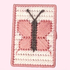 Pode incluir: Uma borboleta rosa com asas cinzas é bordada em uma tela branca. A borboleta é cercada por uma borda rosa.
