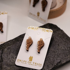 May include: Wooden wing-shaped earrings with intricate carvings, displayed on white cards with gold accents. The cards feature the text "DROPS OF VRAJA TULSI JEWELRY". The earrings are brown and tan, set against a dark gray stone.