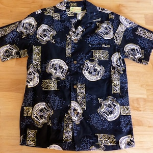 May include: Black short-sleeved shirt with a repeating pattern of shield-like designs in white, grey, and gold. The shirt has a classic collar and a single chest pocket. The fabric appears to be a lightweight material.