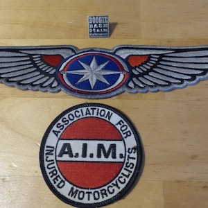May include: Three embroidered patches on a wooden surface. The top patch is a silver wing design with a blue and red emblem. Below is a round patch with the text "ASSOCIATION FOR INJURED MOTORCYCLISTS". A small square patch reads "BOOGIE BASH 94 AL Kelowna".