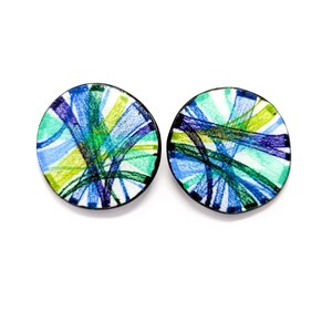 May include: A pair of round earrings with a colorful abstract design. The earrings are made of white enamel with blue, green, and yellow lines. The earrings have a black border.