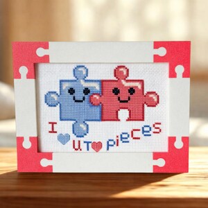 May include: Cross-stitch artwork showing two puzzle pieces, one blue and one red, with smiling faces. The phrase "I U pieces" is stitched below. The artwork is framed in a white and red puzzle-shaped frame.